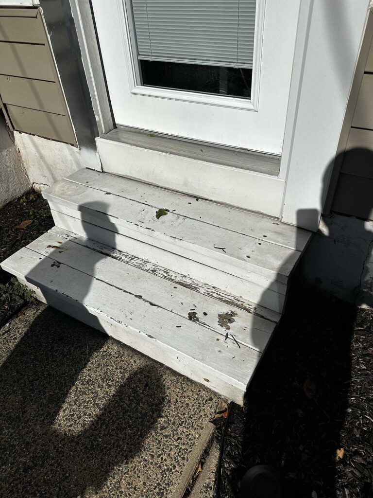 Weathered white-painted wooden steps with peeling paint leading to a door with a window, surrounded by concrete walkway and mulch