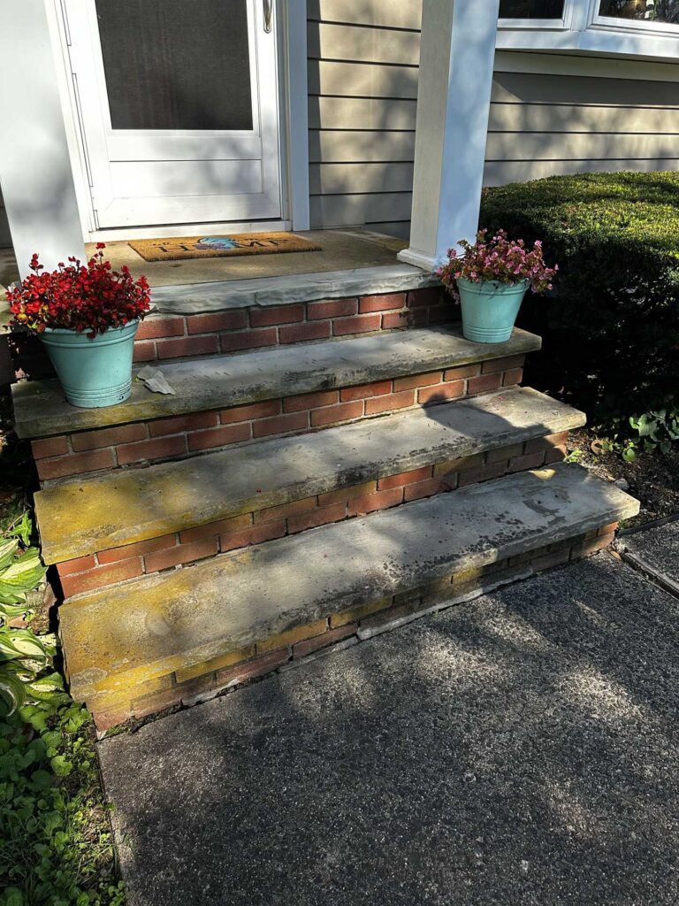 Front entrance with brick and stone steps flanked by two teal flower pots holding red and pink blooms, leading to a screen door with a 'HOME' welcome mat.