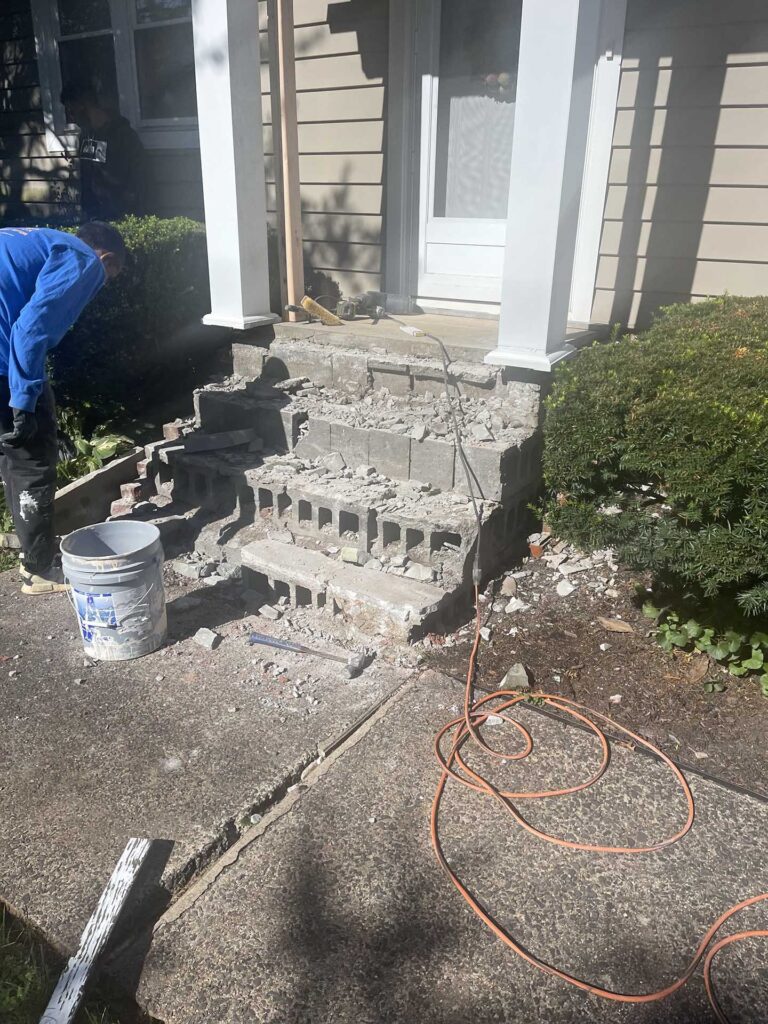 A worker in a blue shirt demolishing old concrete front porch steps, revealing hollow cinder blocks, with rubble, a bucket, hammer, and orange extension cord scattered around the work area.