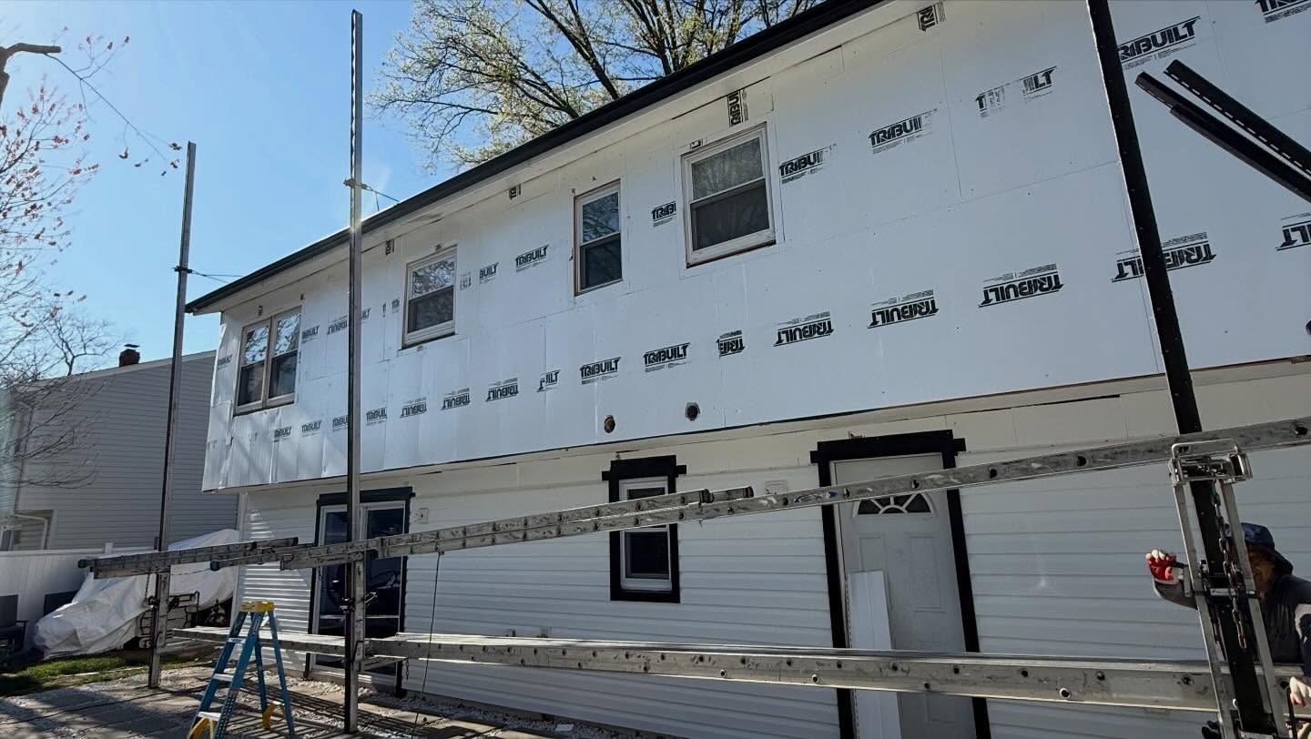 Two-story home mid-project with house wrap exposed above and new white vinyl siding installed on the lower level